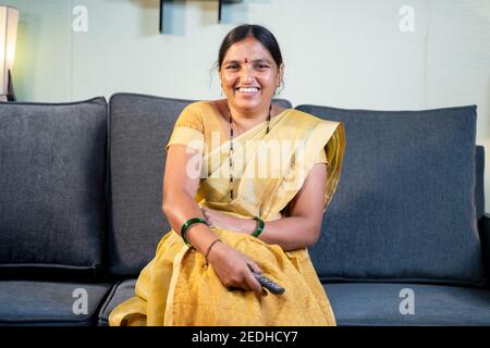 Indian woman in saree watching TV at home while sitting on sofa by holding remote during leisure time at home. Stock Photo