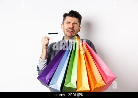 Sad crying man wasted all money on shopping, holding bags and showing empty credit card, standing against white background Stock Photo