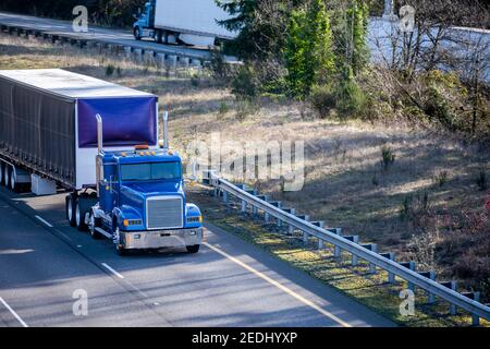 Several different big rigs semi trucks with dry van semi trailers running on the divided highway road with one way traffic lines moving in opposite di Stock Photo