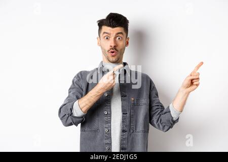 Excited guy showing advertisement aside, pointing fingers right and gasping amazed, checking out special deal, standing on white background Stock Photo