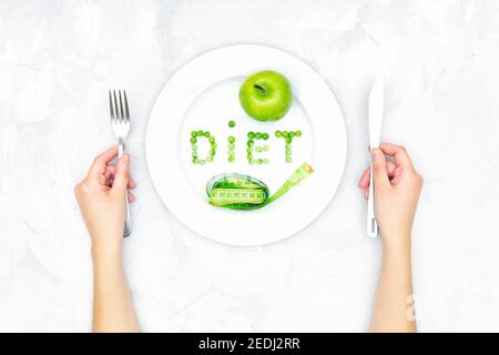 Word DIET arranged of garden peas on a large white plate, a fresh green apple and a soft tape measure on a concrete table. Female hands holding a knif Stock Photo