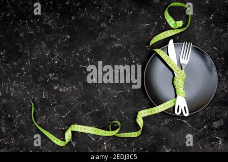 A fork and a knife tied up with a green measuring tape on a black plate on a textured black table. Weight loss concept, copy space. Stock Photo