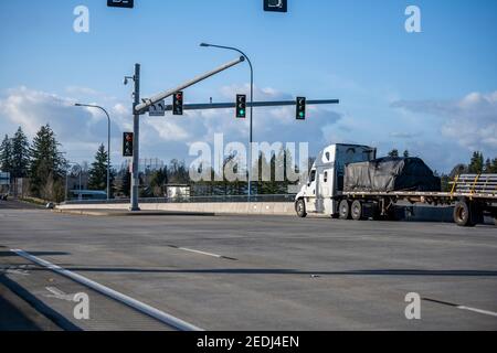 Big rig white semi truck tractor transporting covered commercial cargo on flat bed semi trailer running on the wide city street road with traffic ligh Stock Photo