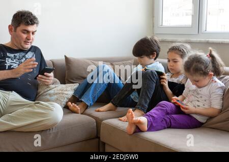 father and his children are sitting on the sofa. Playing internet games. they all have mobile phones and digital tablets in their hands Stock Photo