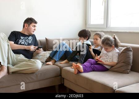 father and his children are sitting on the sofa. Playing internet games. they all have mobile phones and digital tablets in their hands Stock Photo