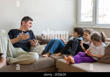 father and his children are sitting on the sofa. Playing internet games. they all have mobile phones and digital tablets in their hands Stock Photo
