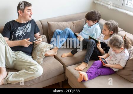 father and his children are sitting on the sofa. Playing internet games. they all have mobile phones and digital tablets in their hands Stock Photo
