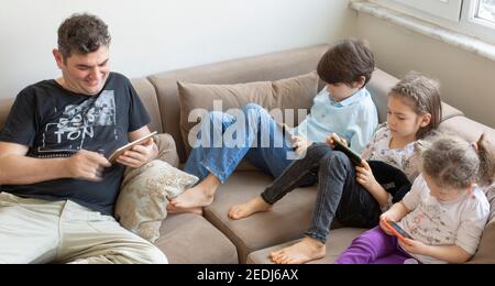 father and his children are sitting on the sofa. Playing internet games. they all have mobile phones and digital tablets in their hands Stock Photo