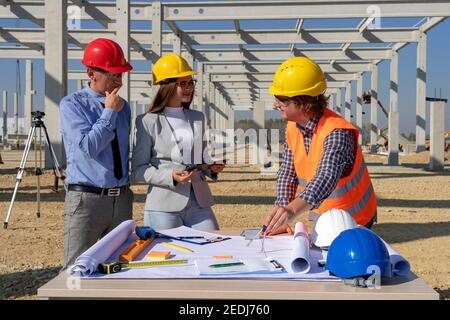 Mixed Construction Team in Yellow and Red Hardhats Discuss on Blueprint on Site Under Construction. Business, Building, Teamwork and People Concept. Stock Photo