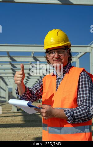 Construction Worker in Yellow Hardhat Standing Under Concrete Construction of a Future Factory. Construction Worker With Clipboard Giving Thumbs Up. Stock Photo