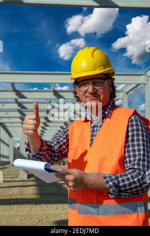 Construction Worker With Clipboard Giving Thumb Up at Construction Site. Happy Worker in Yellow Hardhat Looking at Camera. Stock Photo