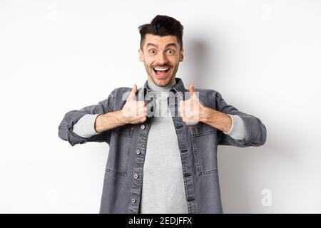 Sounds great. Excited young man showing thumbs up and smiling happy, recommending cool thing, praise nice job, standing on white background Stock Photo