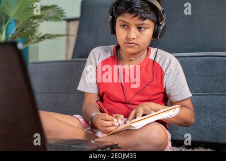 Serious kid with headphone noting down to book by seeing laptop during online class at home - concept of online classroom, online education Stock Photo