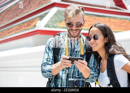 Young interracial tourist couple backpackers looking at smartphone screen finding direction while traveling in Bangkok Thailand Stock Photo