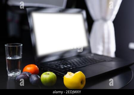 Glass of water and fresh fruits near empty white mock up laptop screen. Healthy snack and working in the office. Bitten apple, orange and plums on Stock Photo
