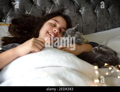 Smiling girls in grey pyjamas lying and slipping on the bed and hugging a cat in the morning Stock Photo
