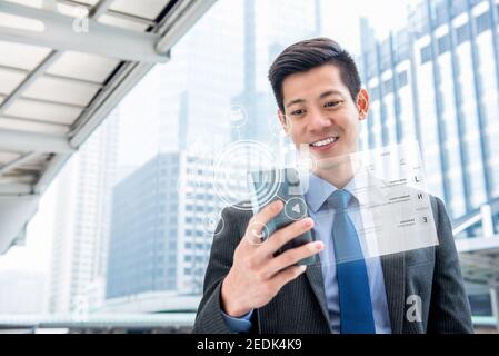 Young handsome Asian businessman using smartphone outdoors with virtual digital interface showing screen and application icons Stock Photo