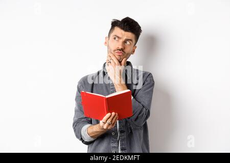 Thoughtful young man plan his schedule, looking aside pensive, holding opened planner or journal, standing on white background Stock Photo