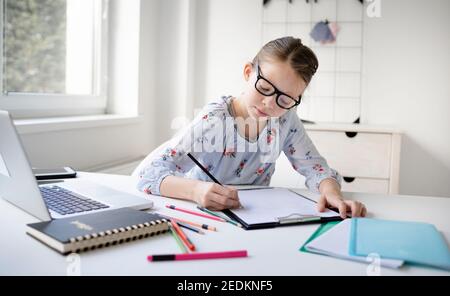 Pretty teenager girl doing homework sitting behind the table at home. Young beautiful girl with glasses, learning online with a laptop, distance learn Stock Photo