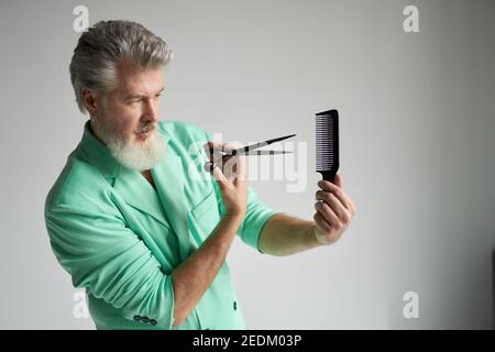 Focused gray haired mature man with beard wearing stylish outfit showing his skills while posing with sharp barber scissors and hair comb over white background Stock Photo