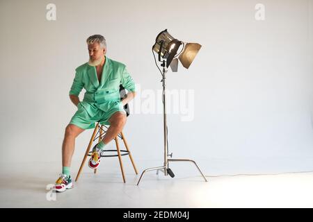 Studio shot of trendy gray haired middle aged man wearing stylish outfit with colorful sneakers looking aside, sitting next to studio spotlight, posing over white background. Fashion, style concept Stock Photo