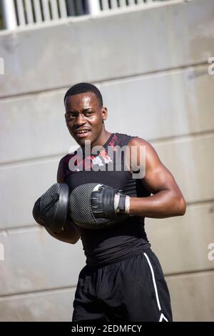 A professional black muscular boxer sweating is putting on his mittens to begin his boxing routine while he smiles at camera. Boxing Concept 2021. Stock Photo