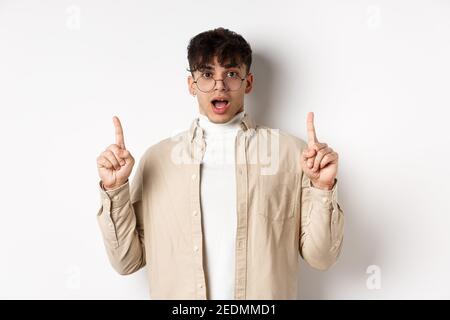 Amazed and excited man in glasses showing awesome promo deal, pointing fingers up and open mouth in awe, standing against white background Stock Photo