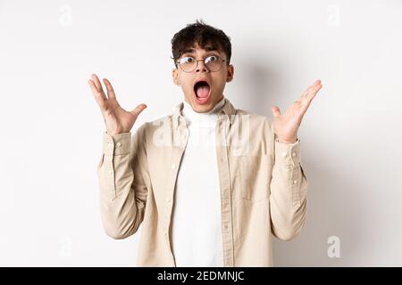 Scared young man in panic, screaming and looking axious, jumping startled and shocked, shaking hands nervously, standing on white background Stock Photo