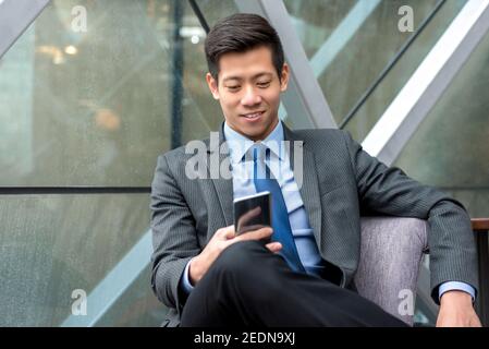 Young handsome Asian Chinese businessman using smartphone searching for information online while sitting at office lounge Stock Photo