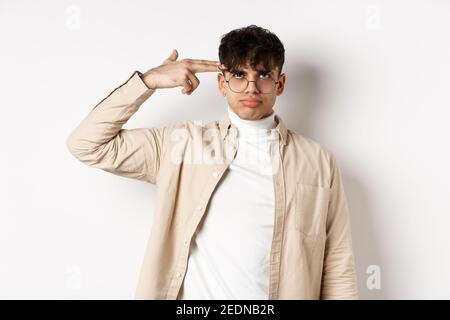 Annoyed young man making finger gun gesture on head, shooting himself from boredom or annoyance, tired of something stupid, standing on white Stock Photo