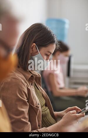 Vertical side view portrait of young woman wearing mask and using smartphone while waiting in line in clinic or office Stock Photo
