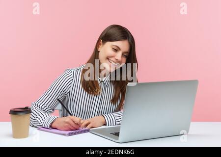 Smiling positive woman office worker in striped shirt making notes with pencil at notepad looking at laptop display, has video call conference. Indoor Stock Photo