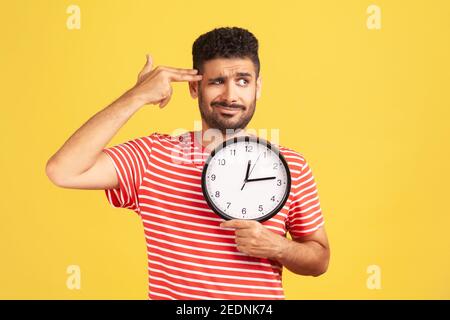 Kill me please. Busy unhappy bearded man in striped t-shirt holding fingers near temple like gun going to make shot holding big wall clock, deadline. Stock Photo