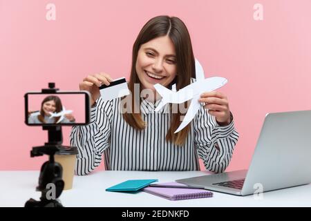 Smiling cheerful woman blogger showing credit card and paper plane on smartphone camera, recording video, giving tips about online booking. Indoor stu Stock Photo