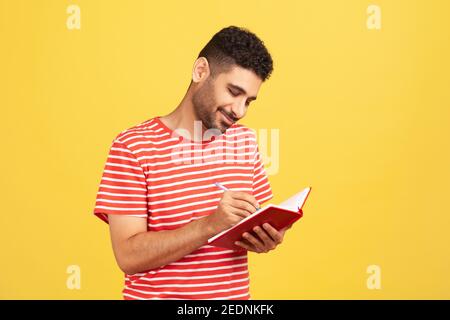 Smiling happy businessman with beard in striped t-shirt writing down to do list, making notes with pen in notebook, checking schedule. Indoor studio s Stock Photo