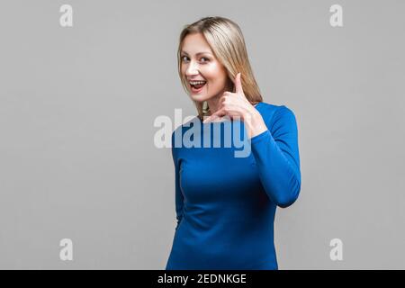 Call me back! Portrait of playful attractive woman in elegant tight blue dress doing talking on phone gesture, smiling and flirting at camera. indoor Stock Photo