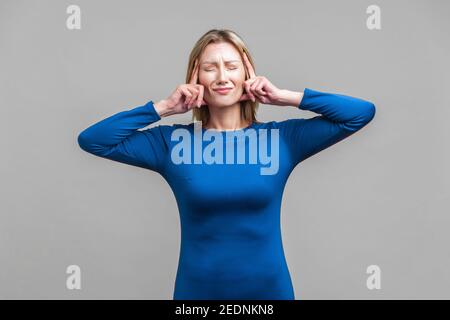 Intense headache. Portrait of upset sick woman in elegant tight blue dress standing touching her temples and grimacing from pain, suffering migraine. Stock Photo