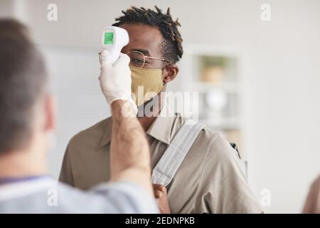 Shot of unrecognizable doctor checking temperature of young African American man wearing mask in line at clinic or hospital, copy space Stock Photo