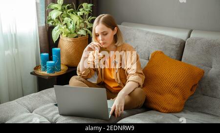 Beautiful young woman shopper makes online purchases using laptop credit debit cards while sitting on couch at home. Woman make urchases thoughtfully. Stock Photo