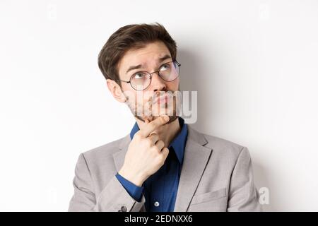 Thoughtful businessman with beard and glasses making decision, looking up and thinking, standing in suit with pensive face, white background Stock Photo