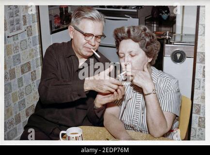 A photographic print from the 1960s shows a middle-aged American man using a disposable gas lighter to light a cigarette for his wife before he lights his own filter-tipped cigarette after the couple had dinner in their small kitchen. Smoking in the United States became increasingly popular in the 19th Century, spurred by the invention of the cigarette rolling machine in 1881, and soon after the introduction of portable “safety” matches. Annual consumption grew from an average of 54 cigarettes smoked by each adult in the country in 1900 to a peak of 4,345 in 1963. Historical photo. Stock Photo
