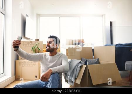 Young man sitting on floor in lounge of new home using mobile phone to make video call with friends and family as he unpacks removal boxes Stock Photo