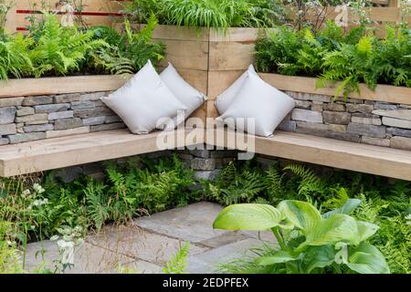 A wooden bench with cushions in a corner seating area and dry stone wall raised beds with ferns and Hosta plants England GB UK Stock Photo