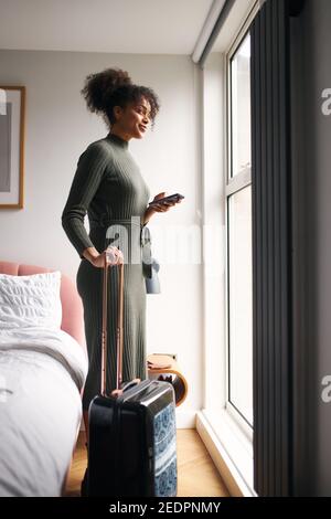 Woman with suitcase checking out of boutique hotel standing by bed waiting for taxi ordered on mobile phone app Stock Photo
