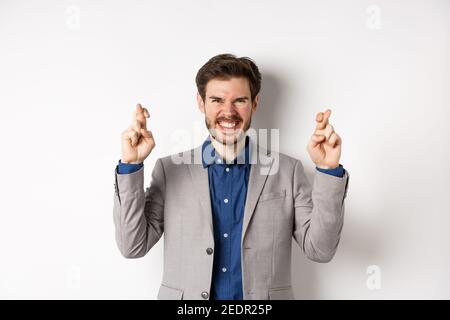 Motivated and excited businessman smiling positive, look hopeful and cross fingers for good luck, making wish, waiting for announcement, white Stock Photo