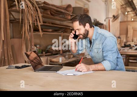 Handyman using phone and laptop computer when scheduling in workshop Stock Photo