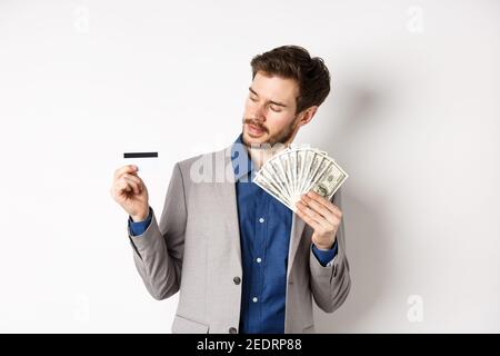 Successful man holding dollar bills and looking at plastic credit card, pleased with bank, standing on white background in business suit Stock Photo