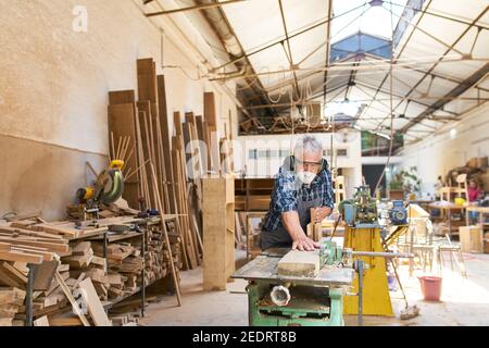 Elderly craftsman in the carpentry workshop while planing wood with a mask and hearing protection Stock Photo