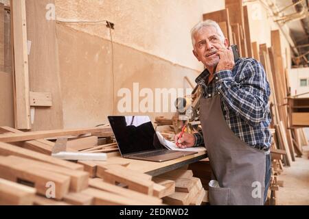 Handyman senior with cell phone and computer when scheduling appointments in the workshop Stock Photo