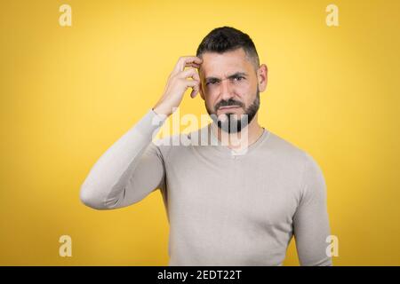 Handsome man with beard wearing sweater over yellow background confuse and wonder about question. Uncertain with doubt, thinking with hand on head Stock Photo